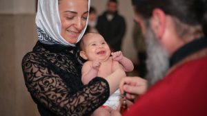 A smiling baby being baptized in a church, held by a woman with a headscarf.