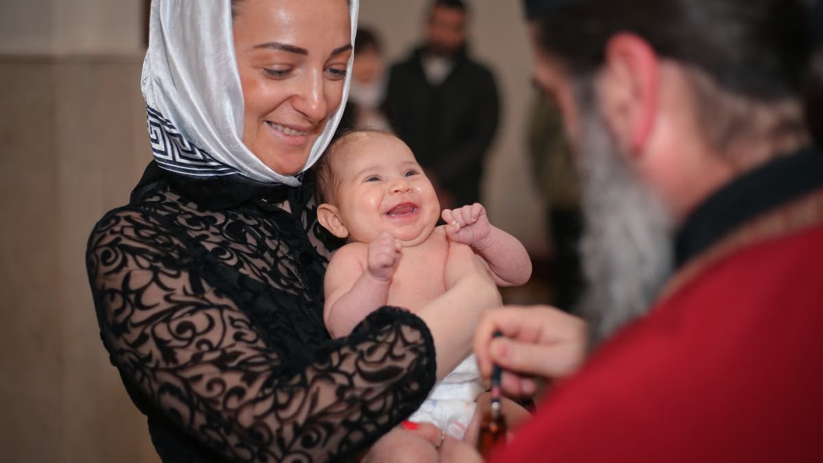 A smiling baby being baptized in a church, held by a woman with a headscarf.